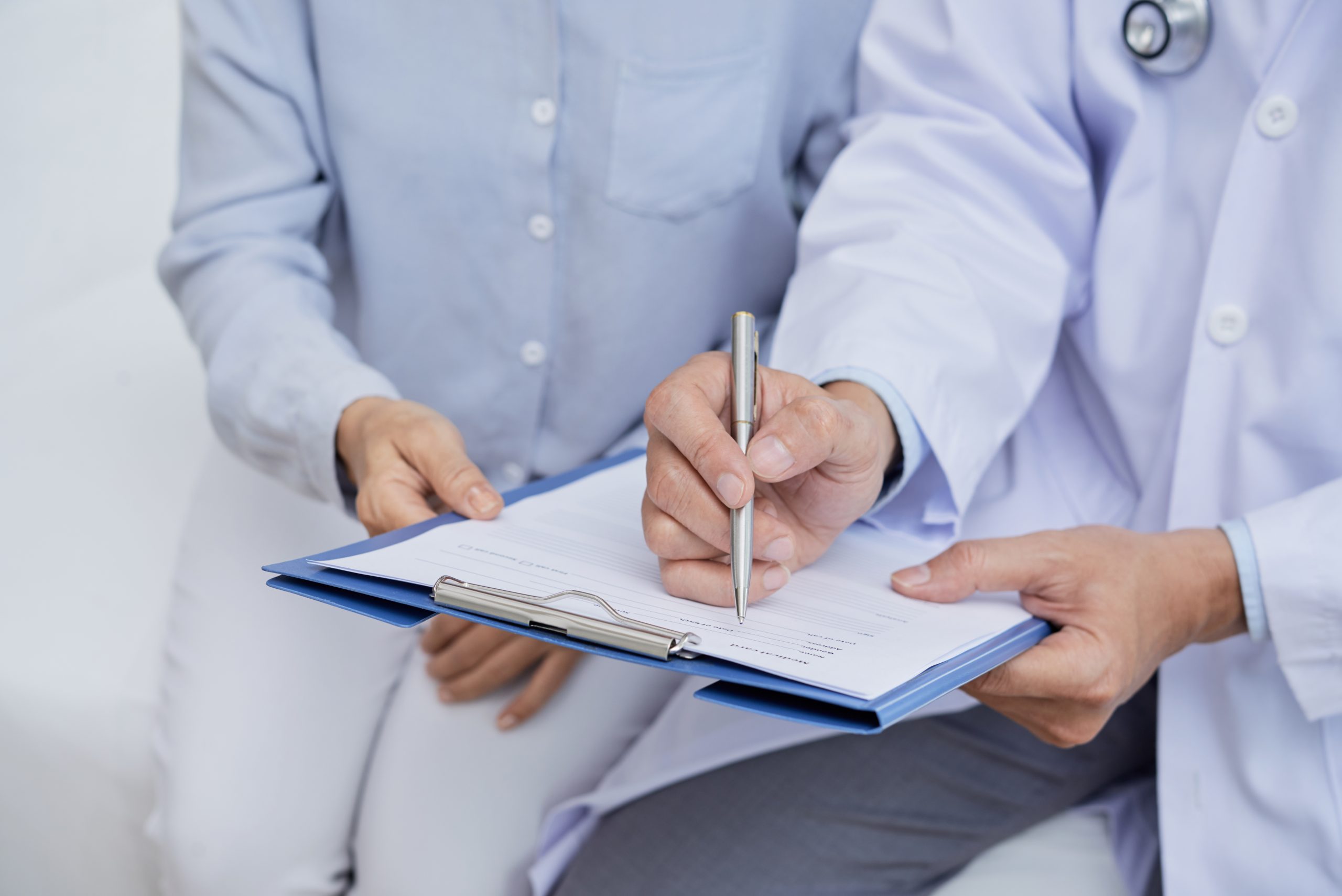 Close-up shot of unrecognizable doctor holding clipboard in hands while helping senior patient to fill in medical form